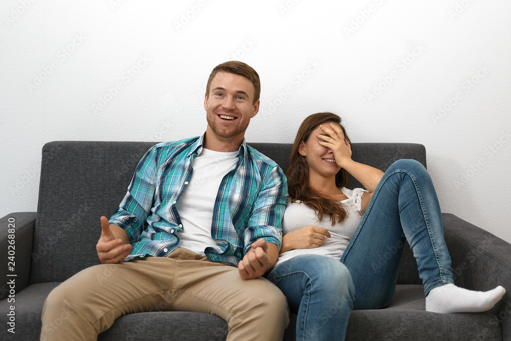 Cute cheerful girl wearing blue jeans covering face and smiling broadly sitting on sofa with her unshaven husband watching TV. Beautiful European couple enjoying nice movie on couch at home
