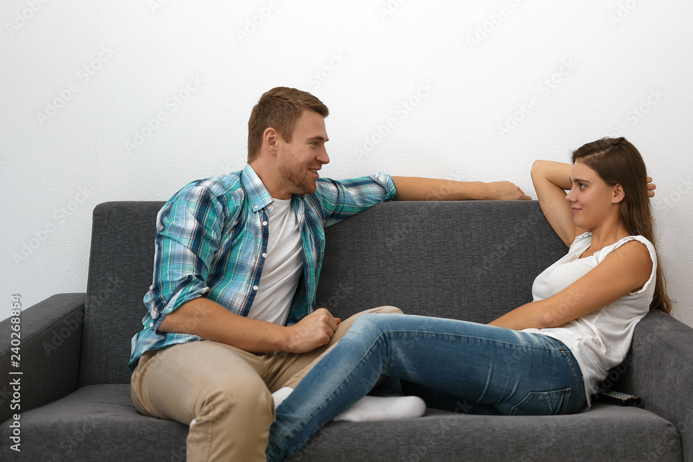 Picture of attractive young woman in t-shirt and jeans sitting comfortably on sofa and listening to handsome bearded man during date, smiling and looking at him with curious facial expression