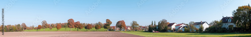 Rural landscape, panoramic view of a small village, with a tractor workingin a field, few houses and many trees with autumn yellow foliage. Bavarian countryside, Germany.