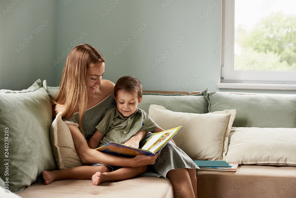 Young mother, read a book to her child, boy in the living room of their home, rays of sun going through the window