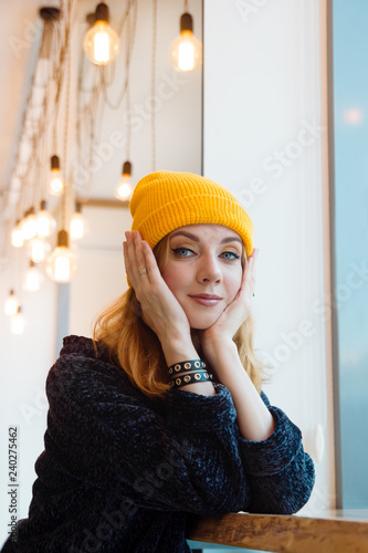 Portrait of young woman with blue eyes and blond hair in a yellow knitting hat in a cafe near to a window.