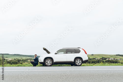 man sitting near broken car with phone, looking for help