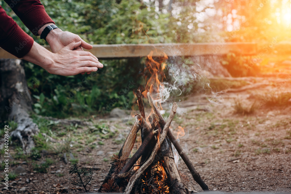 Fototapeta premium Summer camp. Hands man near a campfire at sunset time in the forest . Concept adventure active vacations outdoor.