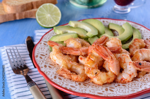 Grilled shrimps next to fresh avocado slices on a plate next to lime, knife and fork on a blue wooden background
