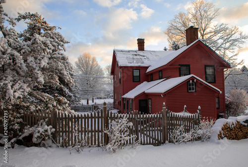 Dark coral painted 18th century New England clapboard home on snowy day at dawn. Scene includes picket fence, snow covered wood pile, snow laden pine trees, and smoke coming from chimney