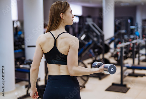 Back view of sporty woman with dumbbell in a modern gym. A strong female is training with dumbbell.