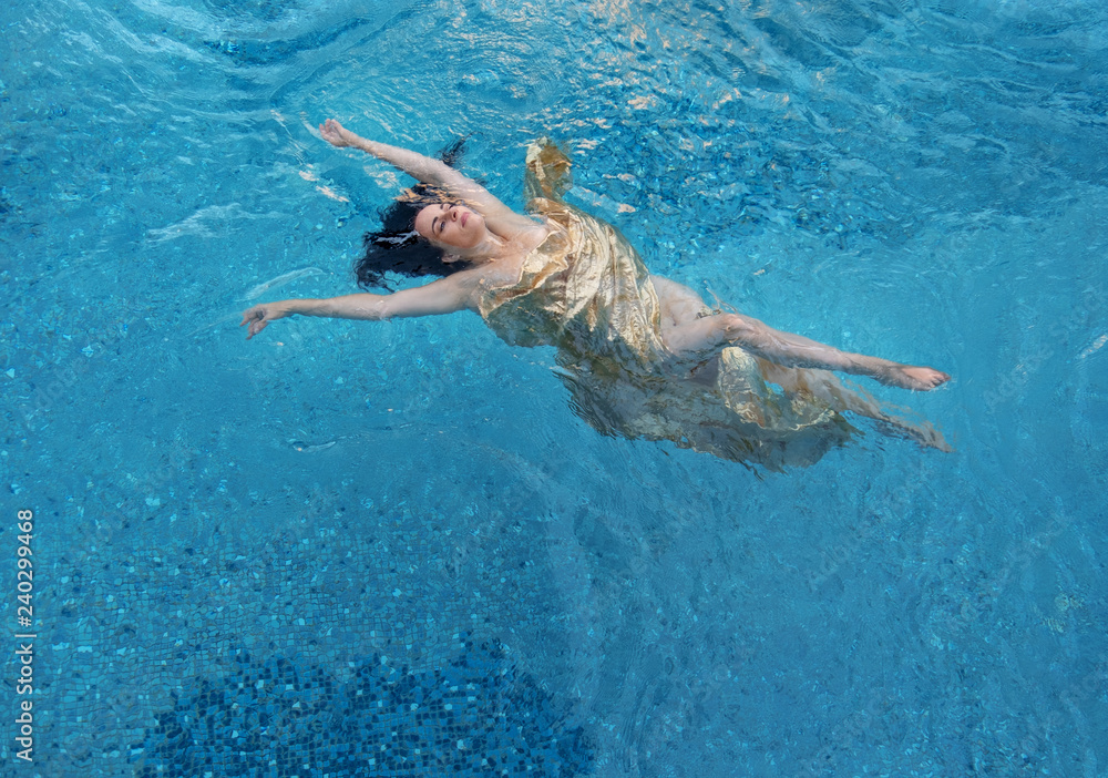 schöne dunkelhaarige Frau mit Locken relaxed in goldenen Tuch schwebt glücklich schwimmend im türkis blauem Wasser im Wellness Pool