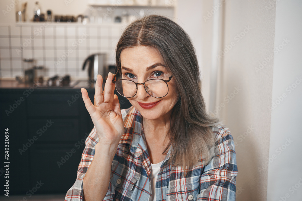Picture of happy charming mature woman wearing checkered shirt and stylish eyeglasses in large round frame, staring at camera with joyful smile, adjusting her eyewear. Optics, vision and age Picture of happy charming mature woman wearing checkered shirt and stylish eyeglasses in large round frame, staring at camera with joyful smile, adjusting her eyewear. Optics, vision and age