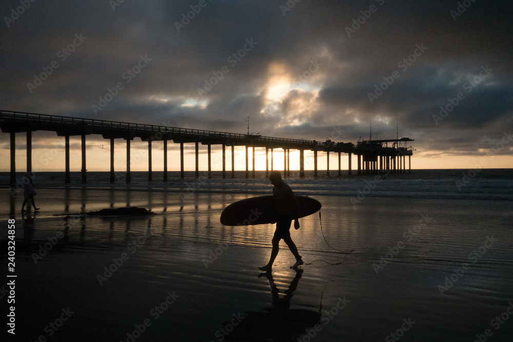 Silhouette of a surfer walking on the beach under a pier in San Diego California