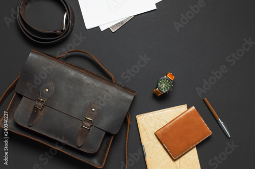 Fashionable concept. Brown leather men's bag, wristwatch, leather passport cover, pen, blank white sheets on black background top view flat lay with copy space. Accessories businessman stylish clothes