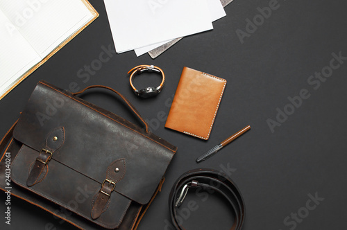 Fashionable concept. Brown leather men's bag, wristwatch, leather passport cover, pen, blank white sheets on black background top view flat lay with copy space. Accessories businessman stylish clothes