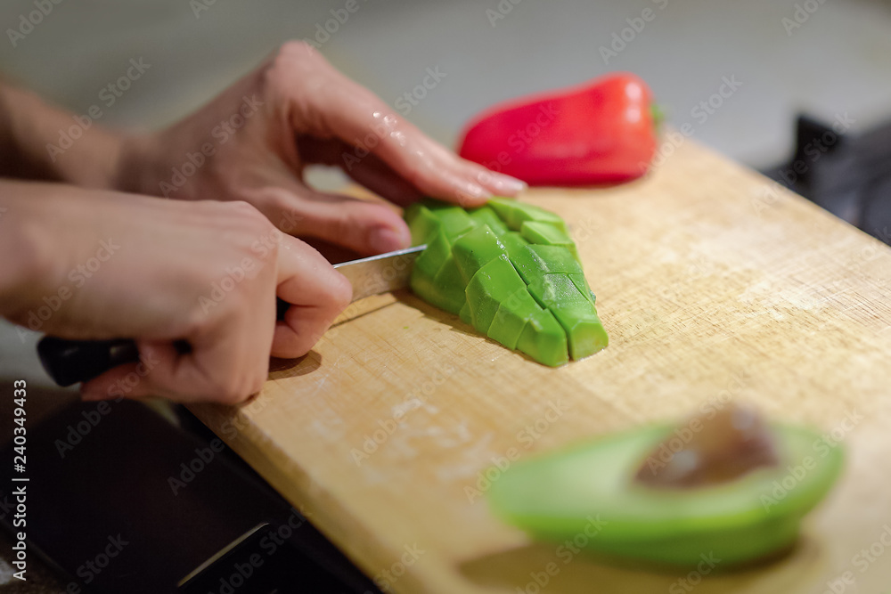 Female hands cutting fresh organic avocado with knife on wooden board in kitchen. Red bell pepper on background. Close up, indoors, copy space. Female hands cutting fresh organic avocado with knife on wooden board in kitchen. Red bell pepper on background. Close up, indoors, copy space.