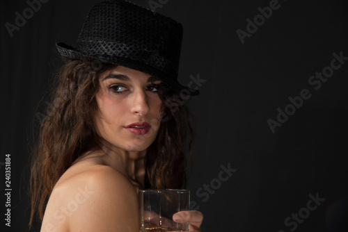 Young girl bare shoulders and black hat on head, with glass in hand. Concept of drinking alcohol the younger generation.Copy space. Black background.