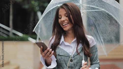 Happy young business woman using smartphone walking on the street in rainy weather, smiling, holding umbrella. Communication concept photo