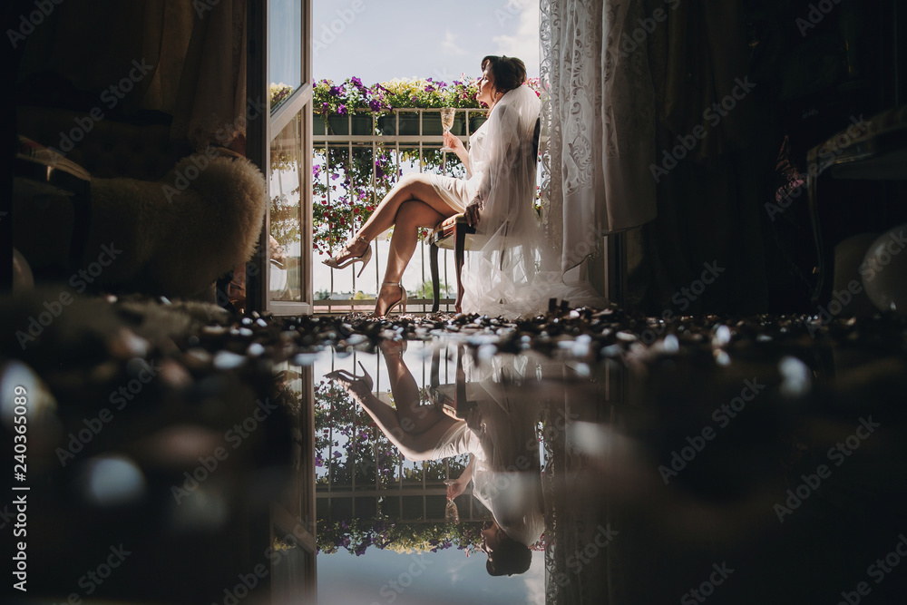 Beautiful stylish brunette bride in silk robe and veil cheering with glass of champagne in the morning on sunny balcony. Sensual portrait of happy woman enjoying wedding. Creative photo Beautiful stylish brunette bride in silk robe and veil cheering with glass of champagne in the morning on sunny balcony. Sensual portrait of happy woman enjoying wedding. Creative photo