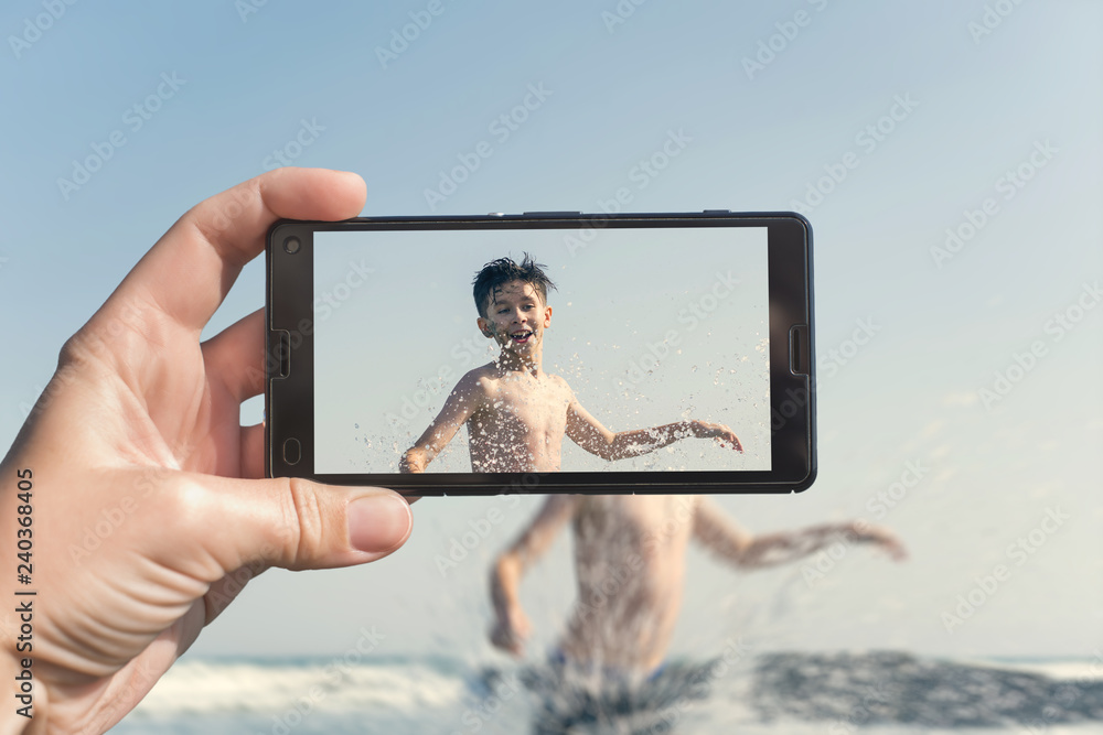 Female taking a picture of a boy on the beach on the phone. Teen boy jumping in sea waves with water splashes. Travel and family concept