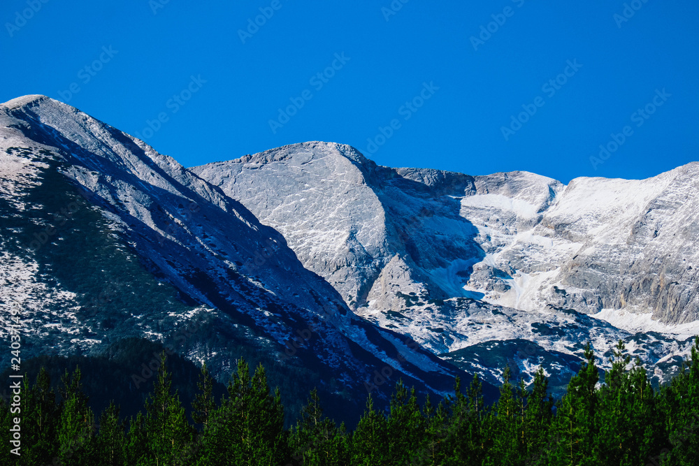 Mountain snow peak, beautiful natural winter backdrop. Ice top of the hill, blue sky background. Alpine landscape, forest, tree line.