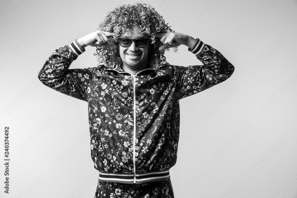 Excited cheerful bizarre man posing om white studio background. Stylish funky male with curly hair black and white portrait. Funny guy in tracksuit expressing feelings. Unusual person in sunglasses.