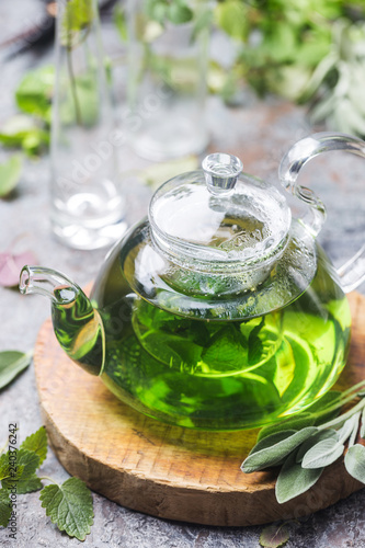 Hot herbal sedative mint tea drink in a glass teapot on wooden tray with fresh garden mint and sage leaves over gray background