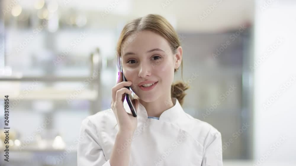 Young lady sous chef looking at camera, smiling chef in chef's form on by professional restaurant kitchen. Young woman is talking on her phone Young lady sous chef looking at camera, smiling chef in chef's form on by professional restaurant kitchen. Young woman is talking on her phone
