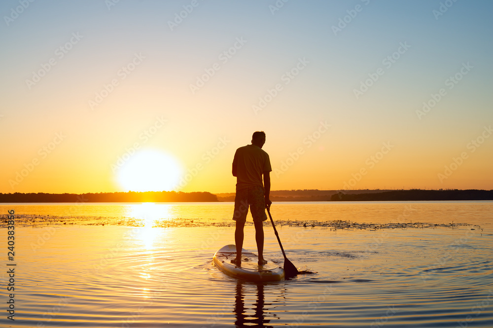 Man is paddling on a SUP board on a large river during sunrise