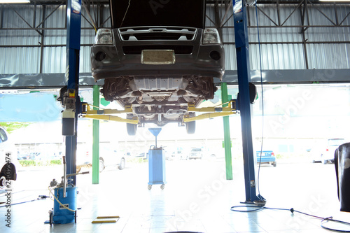 the under view of grey pick up car on lift  at service bay inside the car service center with blue oil tank at bottom and bright daylight from outside