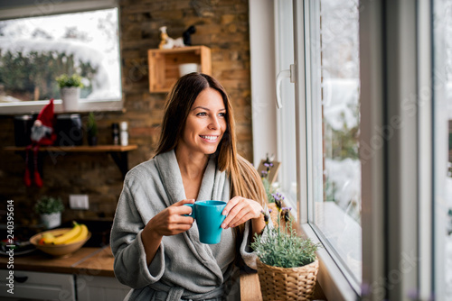 Smiling young woman in bathrobe drinking coffee in the morning, looking through window.
