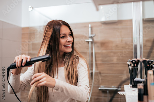 Konstfotografi Beautiful young smiling woman using a hair straightener while looking into the mirror in bathroom