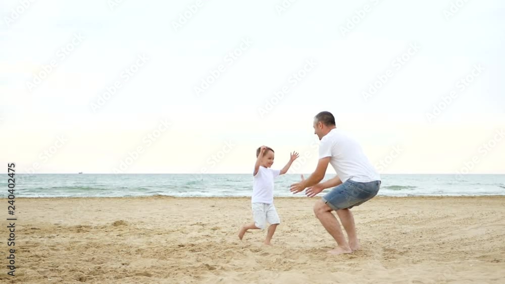 A little joyful boy runs along the sandy seashore to his father. Happy dad plays with the child, raising him in his arms and circling rising to the sky against the background of the sea. A little joyful boy runs along the sandy seashore to his father. Happy dad plays with the child, raising him in his arms and circling rising to the sky against the background of the sea.
