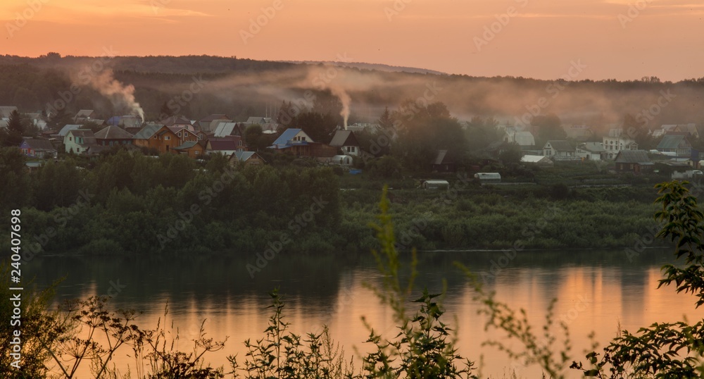 Dawn over the river. Sunrise over the village. Morning rays of the sun. The first rays of the rising sun. The village is at dawn. Smoke from the pipes of houses. Summer sky in the morning. The village