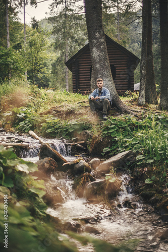 Travel and wanderlust concept. Relaxing with nature. Stylish hipster traveler sitting at river in forest on background of wooden log in mountains. Exploring  woods in summer. Copy space. .