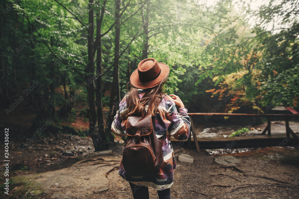 Stylish hipster girl in hat traveling in mountains. Young woman with backpack exploring and walking in summer forest. Travel and wanderlust concept. Space for text. Back view Stylish hipster girl in hat traveling in mountains. Young woman with backpack exploring and walking in summer forest. Travel and wanderlust concept. Space for text. Back view