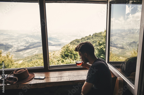 Stylish hipster traveler drinking hot tea in glass cup on background of  beautiful landscape of woods and mountains. View from wooden window on mountain. Space for text. Travel © sonyachny