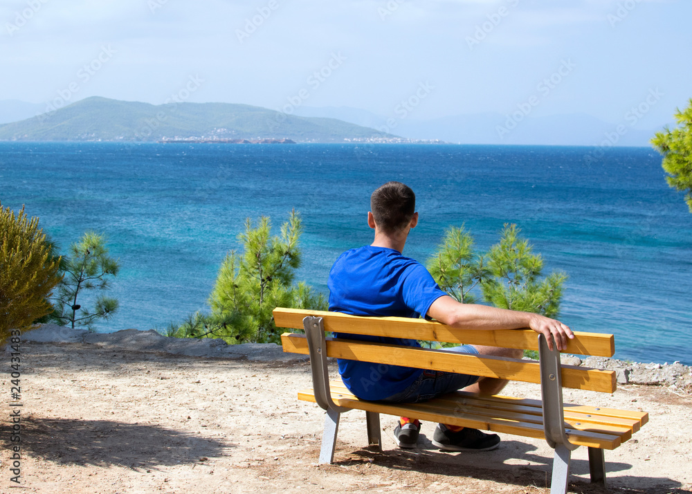 The guy in the blue T-shirt sits alone on a wooden bench on a dais against the background of the Mediterranean Sea in Greece. Sunny summer day, a young guy on a bench on the coast