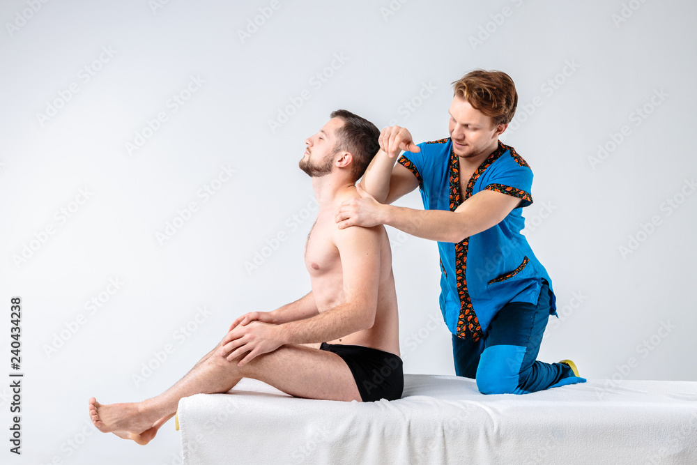 Obraz premium Theme massage and body care. A handsome Caucasian male doctor in blue uniform and beard diagnosing muscles of neck and shoulders, stretching guy with good figure on massage table a white background