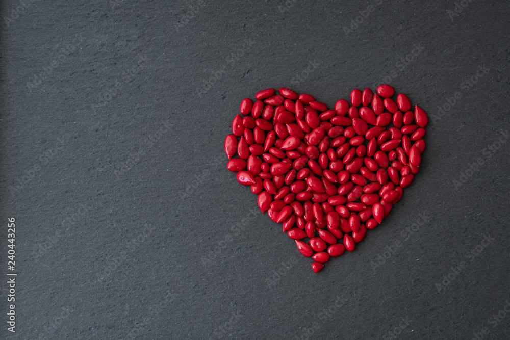 Fototapeta premium Red candy coated sunflower seeds in a heart shape on a gray slate background for Valentine’s Day