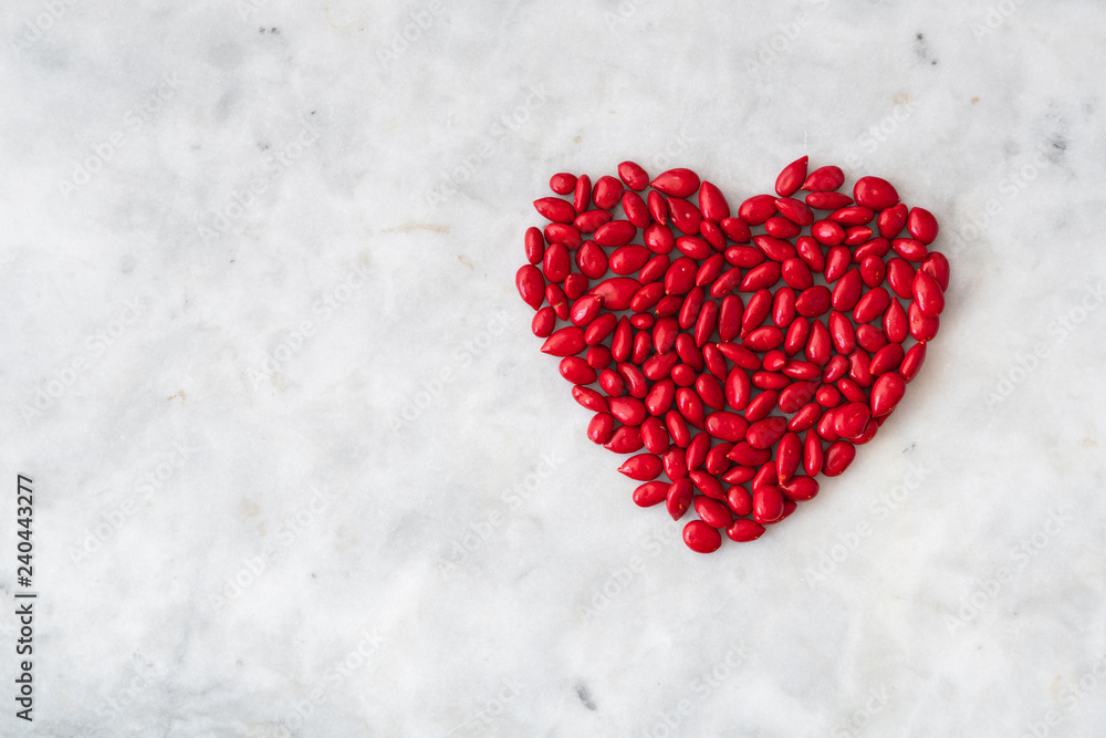 Fototapeta premium Red candy coated sunflower seeds in a heart shape on a white granite background for Valentine’s Day