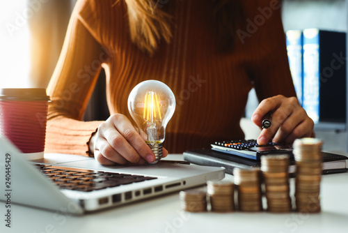business man hand holding lightbulb with using smartphone and calculator to calculate and money stack. idea saving energy and accounting finance in morning light photo