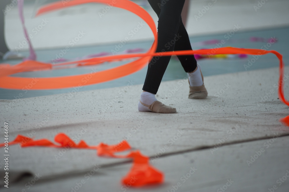 Spirals with orange art ribbon in fitness class. Young little gymnast girl in black sportswear dress, doing rhythmic gymnastics exercise with orange ribbon