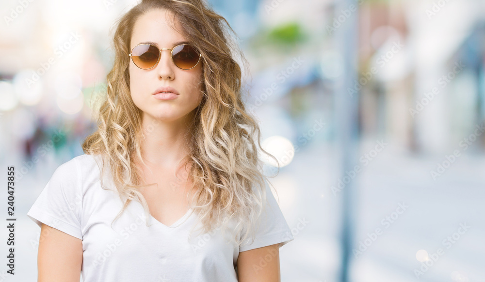 Beautiful young blonde woman wearing sunglasses over isolated background with serious expression on face. Simple and natural looking at the camera. Beautiful young blonde woman wearing sunglasses over isolated background with serious expression on face. Simple and natural looking at the camera.