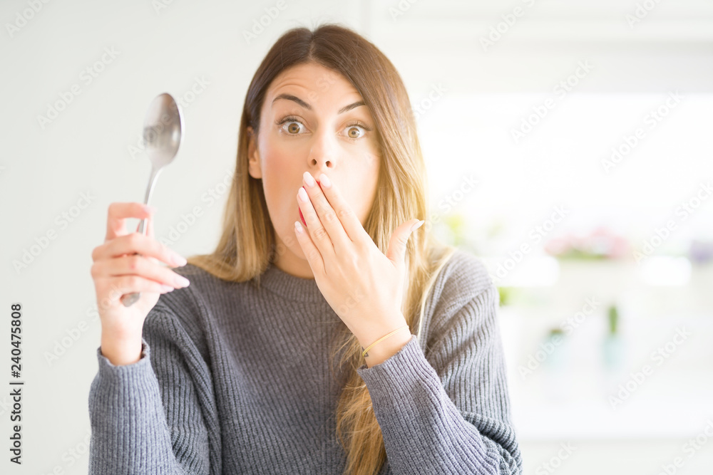 Young beautiful woman holding silver spoon at home cover mouth with hand shocked with shame for mistake, expression of fear, scared in silence, secret concept Young beautiful woman holding silver spoon at home cover mouth with hand shocked with shame for mistake, expression of fear, scared in silence, secret concept
