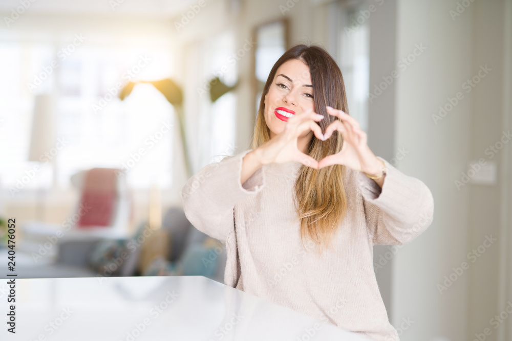 Young beautiful woman wearing winter sweater at home smiling in love showing heart symbol and shape with hands. Romantic concept. Young beautiful woman wearing winter sweater at home smiling in love showing heart symbol and shape with hands. Romantic concept.