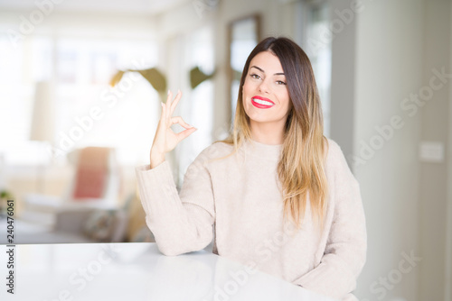 Young beautiful woman wearing winter sweater at home smiling positive doing ok sign with hand and fingers. Successful expression.