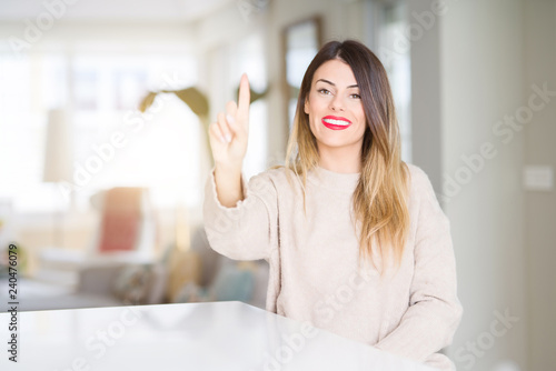 Young beautiful woman wearing winter sweater at home showing and pointing up with finger number one while smiling confident and happy.