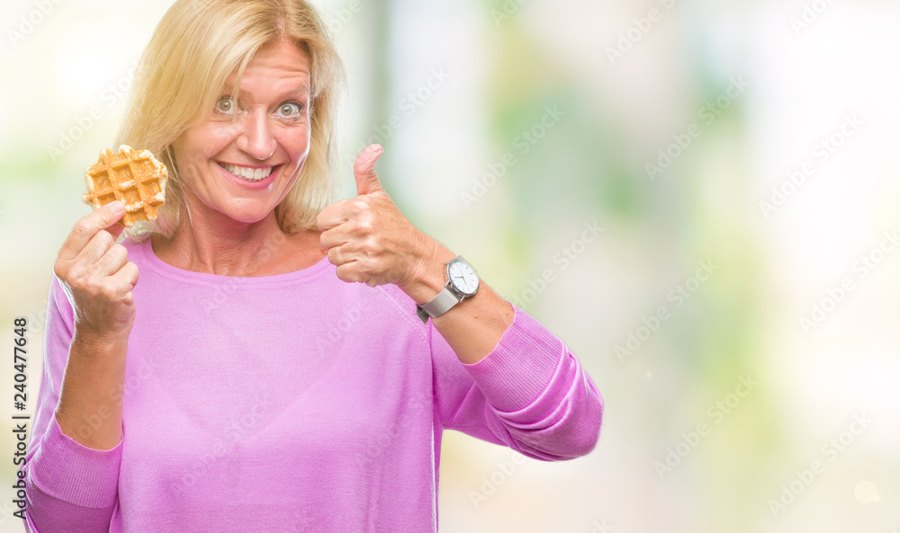 Fototapeta premium Middle age blonde woman eating sweet waffle over isolated background happy with big smile doing ok sign, thumb up with fingers, excellent sign