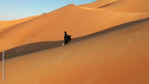  Aerial view from a drone flying next to a woman in abaya (United Arab Emirates traditional dress) walking on the dunes in the desert. Dubai, UAE.