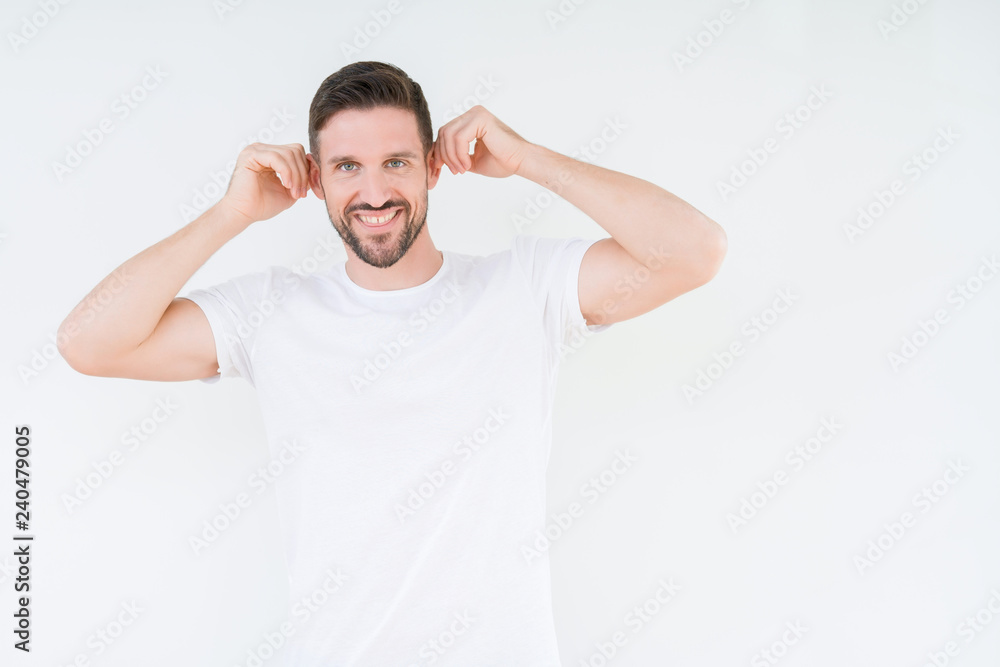 Young handsome man wearing casual white t-shirt over isolated background Smiling pulling ears with fingers, funny gesture. Audition problem Young handsome man wearing casual white t-shirt over isolated background Smiling pulling ears with fingers, funny gesture. Audition problem