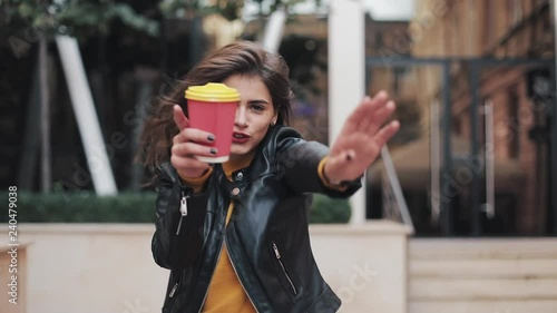 Happy young woman walking near the business center and dancing with a cup of coffee in hand. Beautiful girl looking into the camera. Good mood, funny, slow motion photo