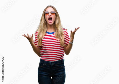 Young beautiful blonde woman wearing sunglasses over isolated background crazy and mad shouting and yelling with aggressive expression and arms raised. Frustration concept.