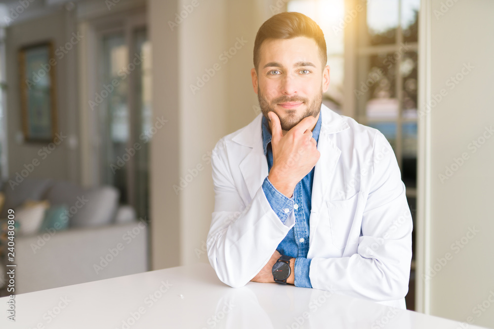Young handsome doctor man at the clinic looking confident at the camera with smile with crossed arms and hand raised on chin. Thinking positive.